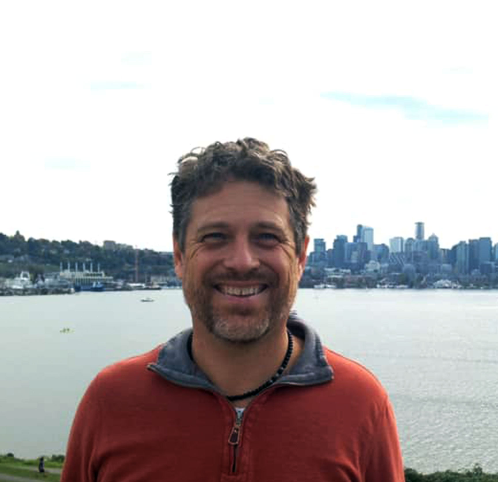 Howard Lamb of Sunergy Systems, championing solar innovation and policy leadership in the Pacific Northwest, smiling outdoors with city skyline and waterfront in the background.